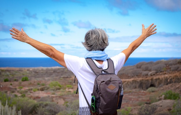 Rear view of active senior woman enjoying freedom and outdoor trekking standing with outstretched arms looking at the horizon over sea