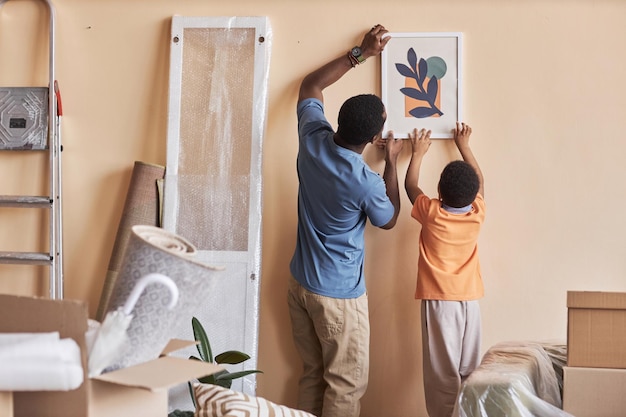 Photo rear view of african american man and his little son hanging picture on wall