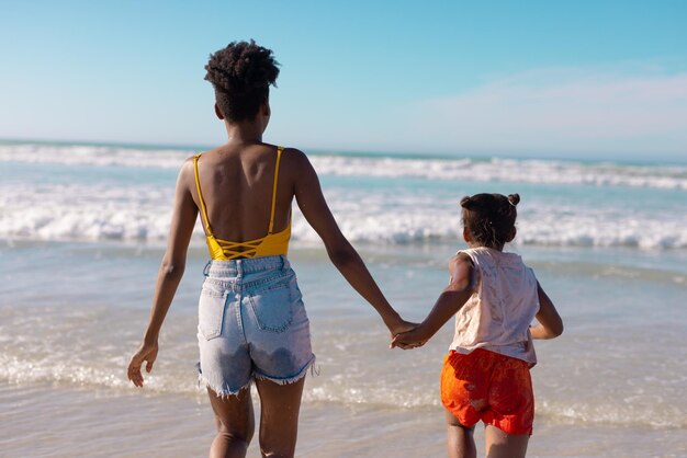 Rear view of african american young woman holding daughter's hands and running in sea against sky