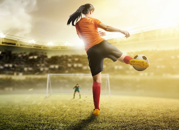 Rear view of asian football player woman in orange jersey kicking the ball in the penalty box