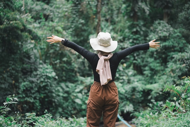 Rear view of asian muslim hiker woman brown hijab standing in front of the tropical forest with her hands raised.