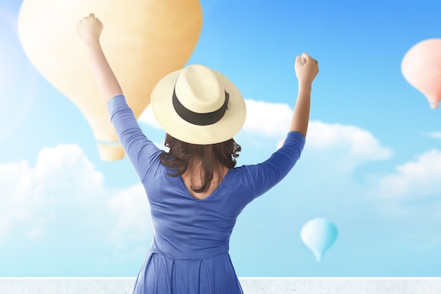 Вид сзади азиатской женщины в шляпе, смотрящей на красочный воздушный шар, летящий на фоне голубого неба
