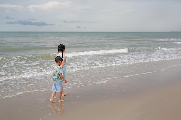 Foto vista posteriore la giovane sorella asiatica e il fratellino camminano insieme sulla spiaggia di sabbia tropicale all'alba il ragazzo e la ragazza di famiglia felici si divertono durante le vacanze estive