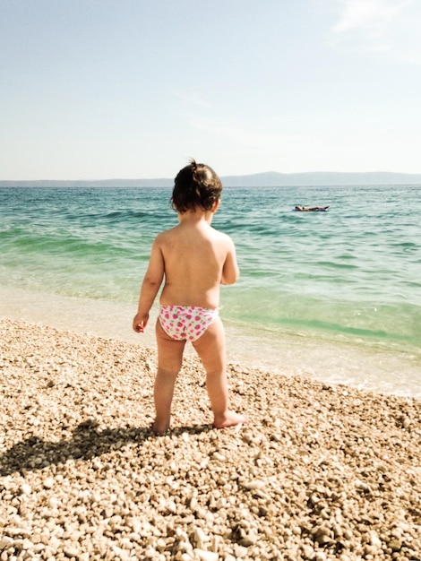 Rear view of baby girl standing at sea shore against sky