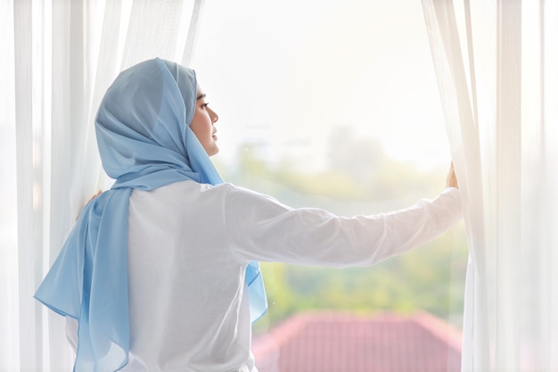 Rear view beautiful asian muslim woman wearing white sleepwear, stretching her arms after getting up in the morning at sunrise. Cute young girl with blue hijab standing and relaxing while looking away