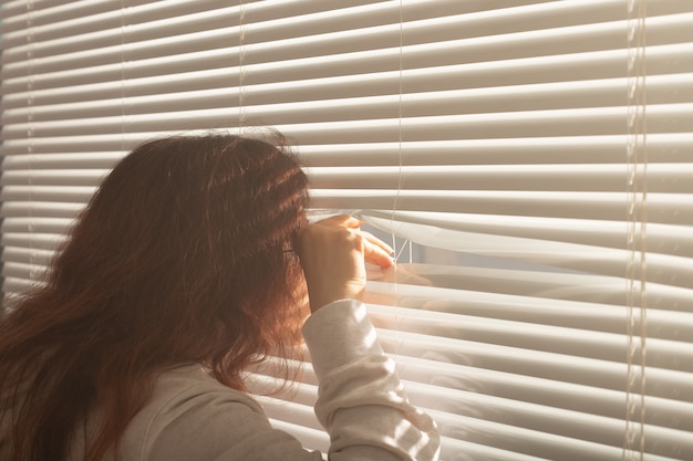 Rear view of beautiful young woman with long hair peeks through hole in the window blinds and looks out the window. Surveillance and curiosity concept