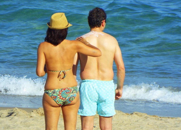 Rear view of bikini woman applying suntan lotion on boyfriend at beach