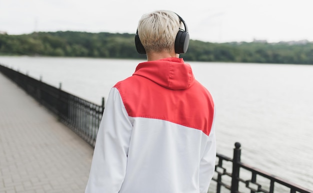 Vista posteriore del giovane ragazzo caucasico biondo che cammina nel parco guardando il lago e ascoltando musica all'aperto concetto di stile di vita e tecnologia della gente