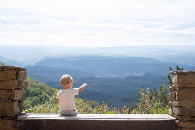 ベンチに屋外に座って、美しい自然の景色を楽しんでいるリアビュー金髪の子供の男の子