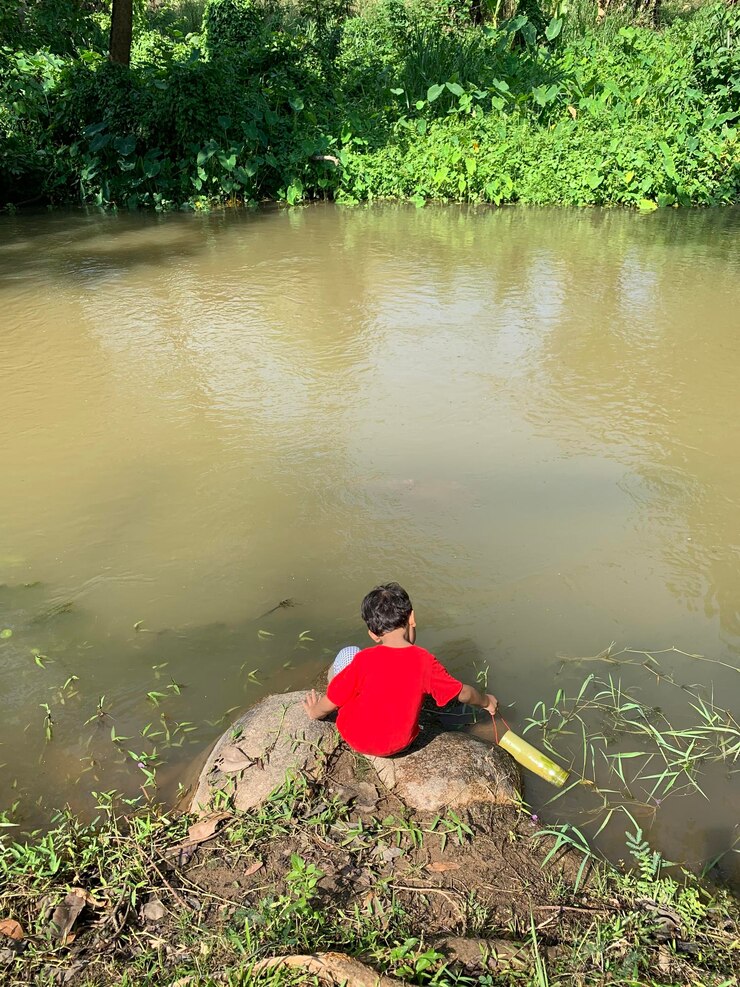 फोटो rear view of boy filling water in container while crouching by river against trees
