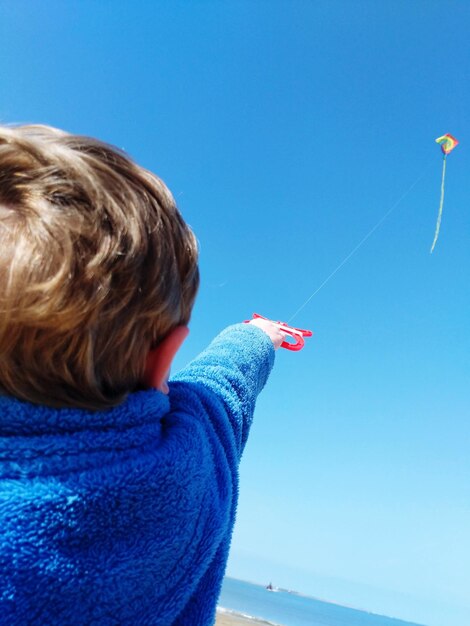 Rear view of boy flying kite against blue sky