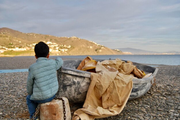 Rear view of boy sitting on rock next to an abandoned boat on the beach