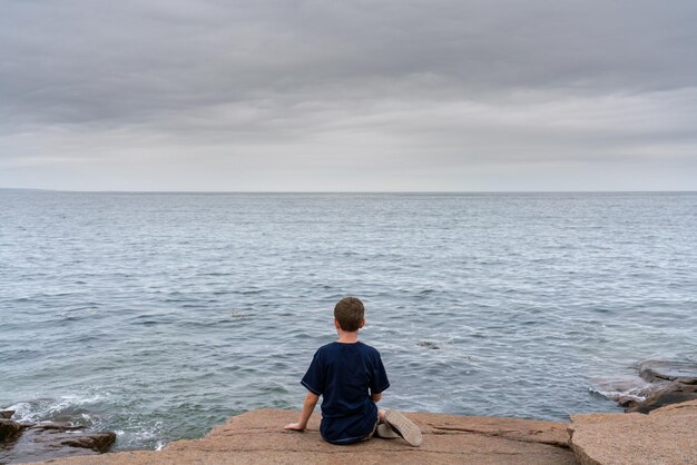 rear view of boy sitting at sea shore against sky