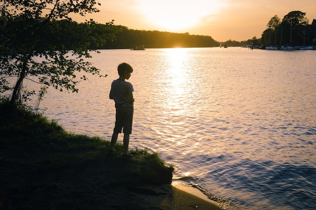 夕暮れ の 時 に 湖 の 横 に 立っ て いる 少年 の 後ろ の 景色