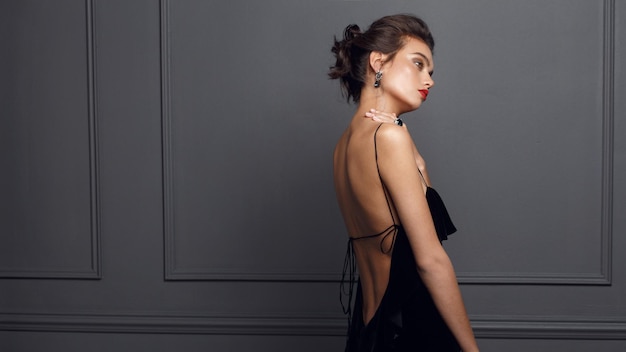 Rear view of a brunette woman in sexy black dress with bare back and beautiful earrings near grey dark wall.