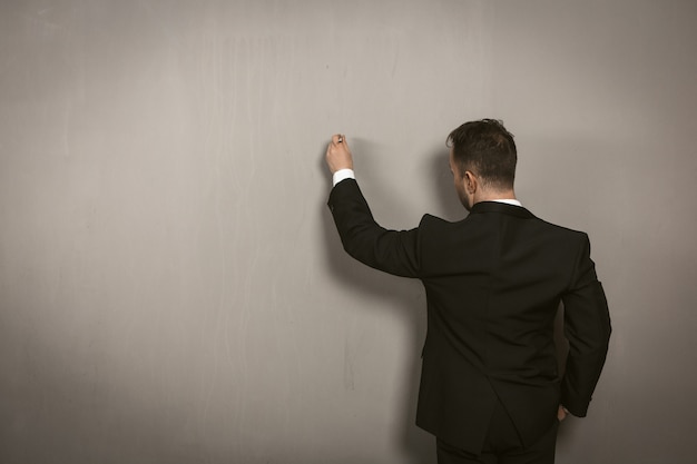 Rear view of businessman writing on wall. Creative man in black elegance suit drawing by pan or chalk on light grey wall. Template concept. Toned image