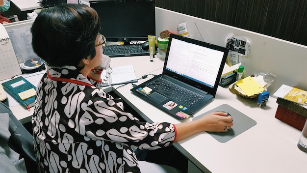 Photo rear view of businesswoman talking on phone while using laptop in office