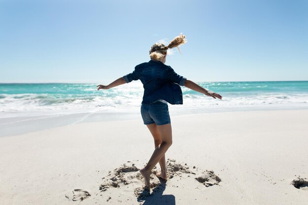 Rear view of a Caucasian girl on a sunny beach, dancing barefoot in the sand with blue sky and sea in the background
