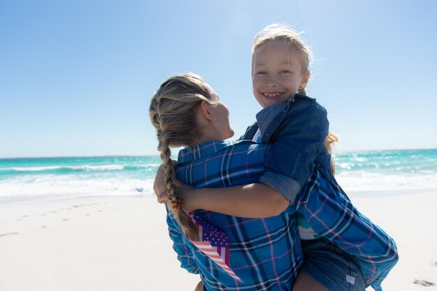 Rear view of a Caucasian girl with her mother, standing on the beach with blue sky and sea in the background, embracing and smiling to camera