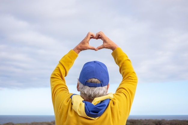 Rear view of Caucasian man standing in front of the sea with the Ukrainian flag around her neck making heart shape with hands as message of love for the Ukrainian people No war freedom and peace