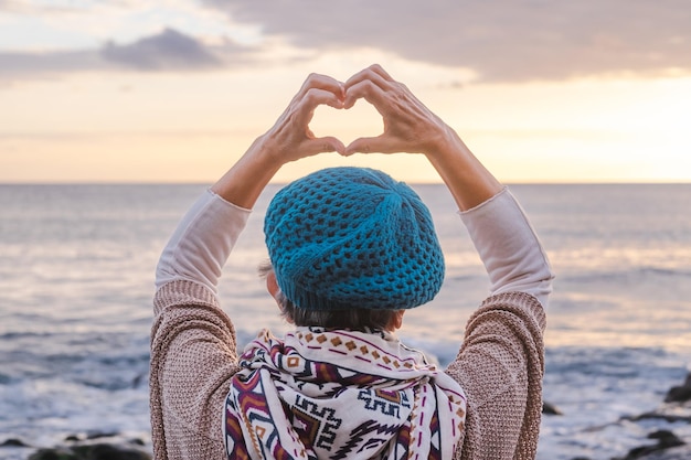 Rear view of caucasian senior woman in front to the sea at\
sunset light admiring horizon over water making heart shape with\
hands elderly romantic lady enjoying nature and freedom