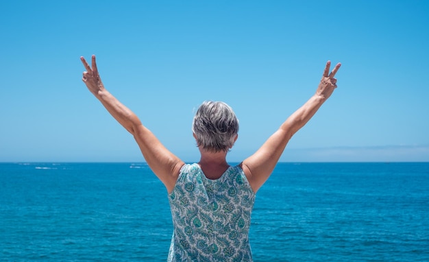 Rear view of caucasian senior woman in front to the sea with
outstretched arms gesturing winner sign with hands feeling freedom
looking at the horizon copy space