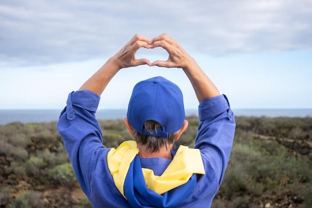 Vista posteriore della donna caucasica in piedi di fronte al mare con la bandiera ucraina intorno al collo che fa a forma di cuore con le mani come messaggio d'amore per il popolo ucraino nessuna guerra, libertà e pace