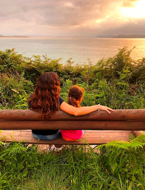 Photo rear view of couple mother and daughter sitting on a bench by sea against sky
