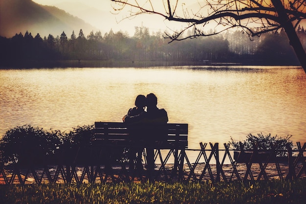 rear view of couple sitting on bench at lakeshore during sunset