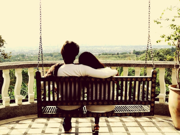 Photo rear view of couple sitting on swing in balcony against clear sky