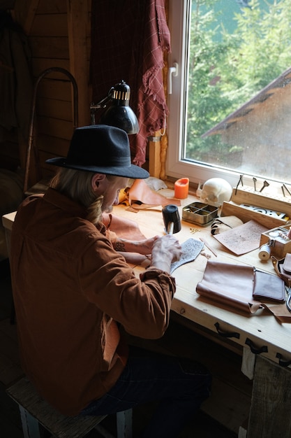 Rear view of craftsman sitting at his workplace and working with hammer, he making products from leather textile