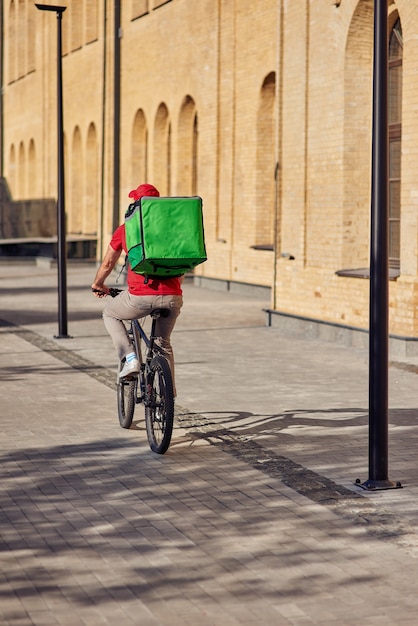 Foto vista posteriore del fattorino con borsa del frigorifero in bicicletta lungo la strada piastrellata al giorno di sole. concetto di consegna del cibo