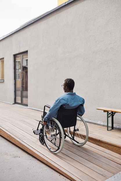 Rear view of disabled young African-American man sitting in wheelchair and moving along ramp near house