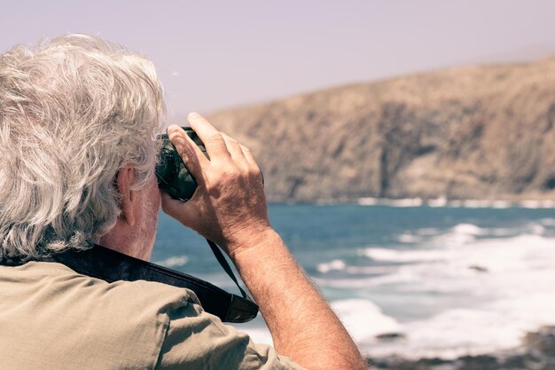 Vista posteriore dell'uomo anziano che tiene il binocolo guardando il mare, la montagna. anziano in pensione che gode di libertà e conoscenza