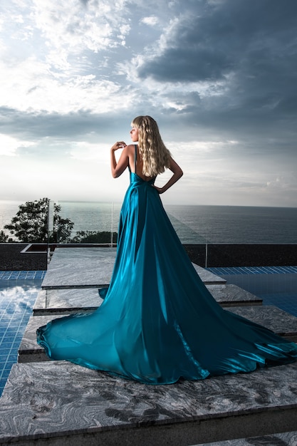 Vista posteriore: elegante bionda in posa in un lungo abito da sera blu vicino alla piscina a sfioro. rilassamento lussuoso. vista sul mare.