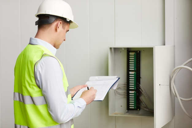 Rear view of engineer in reflective vest making notes in document he checking the data of electricity meter