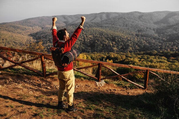 Rear view of explorer having hands raised. It's a beautiful sunny autumn day. Man having backpack and blanket on backs. Freedom.