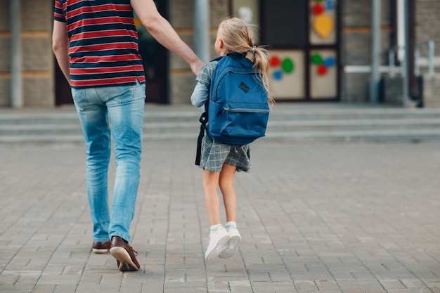 Rear view of father walking back to school with his daughter carrying backpack.