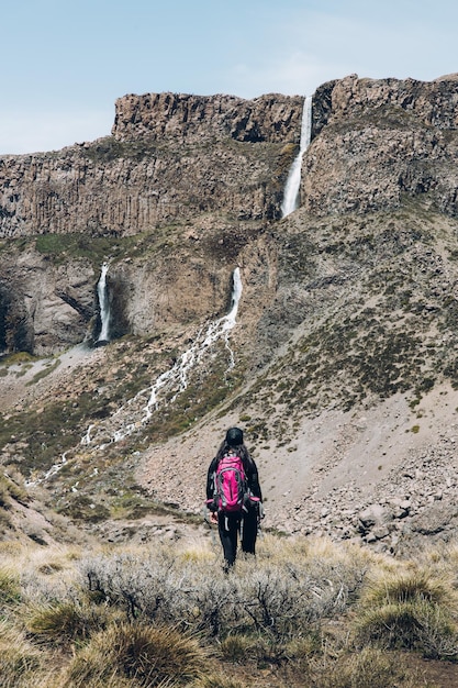 Rear view of female backpacker hiking through a rocky path to a big waterfall. Vertical shot