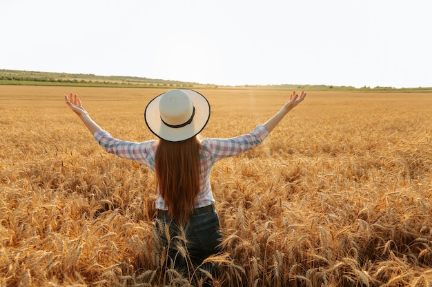 Rear view of female farmer with hat on head in golden wheat field at sunset harvest time sunny summer day