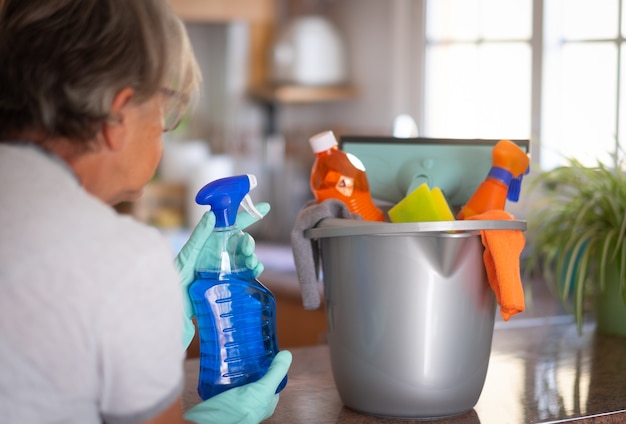 Rear view of a female people ready for household chores while holding a gray plastic bucket with cleaning items in her hands. Bright light from the window