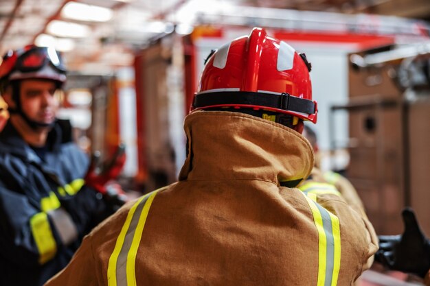 Rear view of firefighter putting on his protective clothes na d helmet while standing in fire brigade.