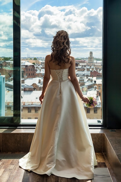 Rear view french bride with bouquet in white dress at background of urban window From behind bridal portrait of pretty fiancee indoors