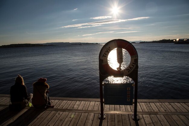 Photo rear view of friends sitting by life belt on pier over lake against sky