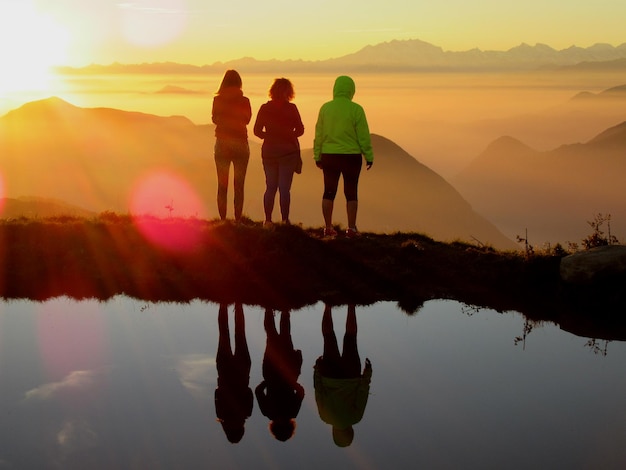 Rear view of friends standing by pond while looking at mountains during foggy weather