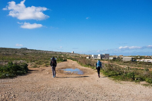 Rear view of friends walking on dirt road against blue sky