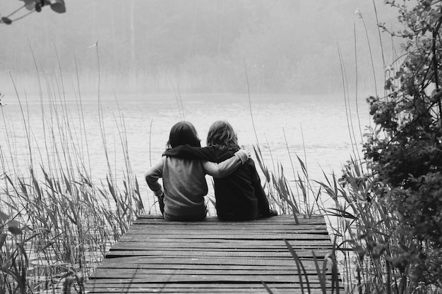 Photo rear view of friends with arm around sitting on pier by river during foggy weather