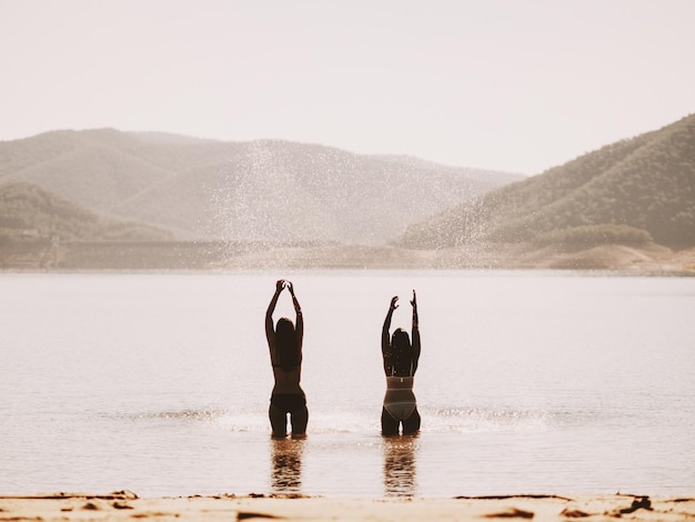 rear view of friends with arms outstretched standing on beach by mountains against clear sky