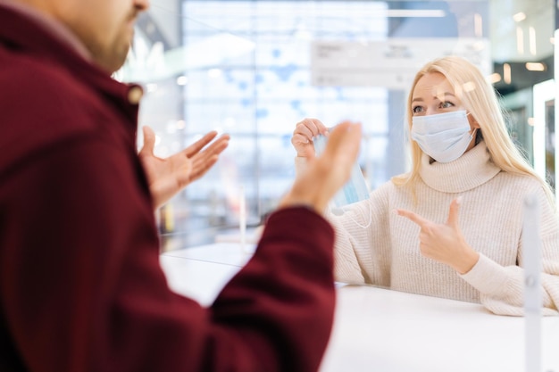 Rear view from shoulder of unrecognizable baffled man talking through glass partition with blonde female staff in face mask pointing to medical protective mask