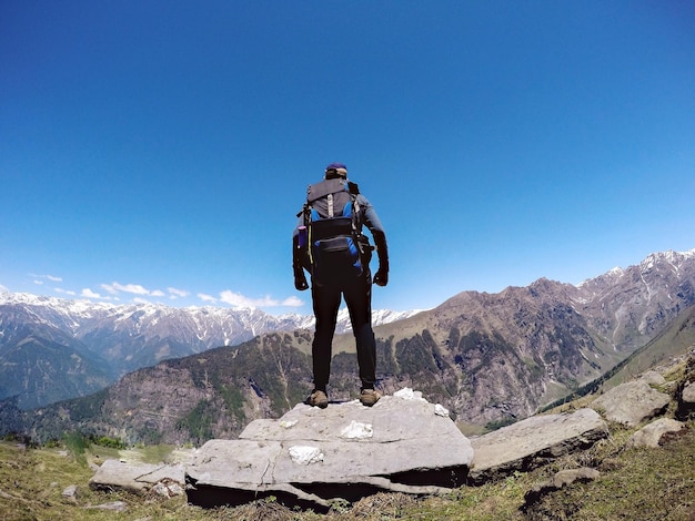明るい青い空を背景に山の岩の上に立っているハイカーの全長の後ろの景色